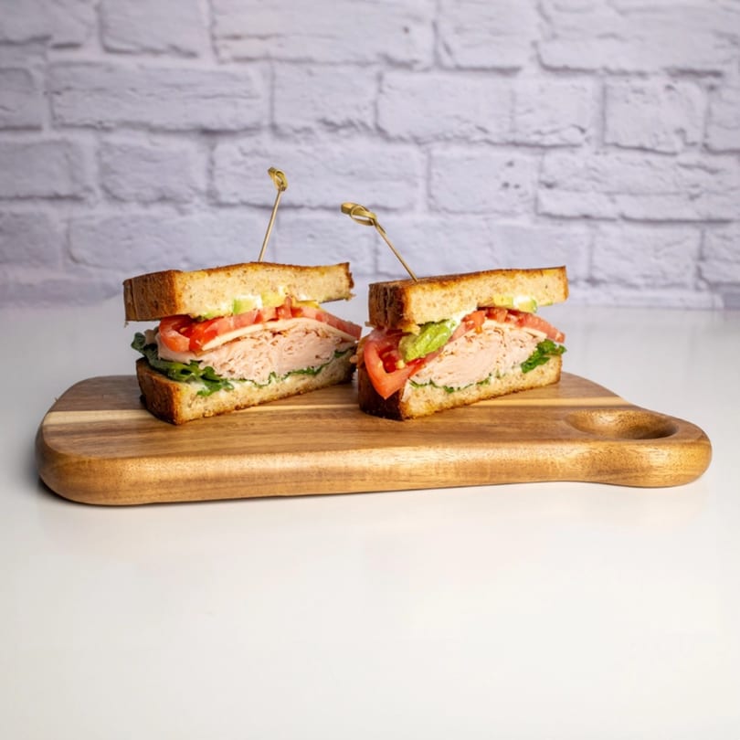 Two halves of a turkey club sandwich with avocado, tomato, and lettuce on a wooden board.