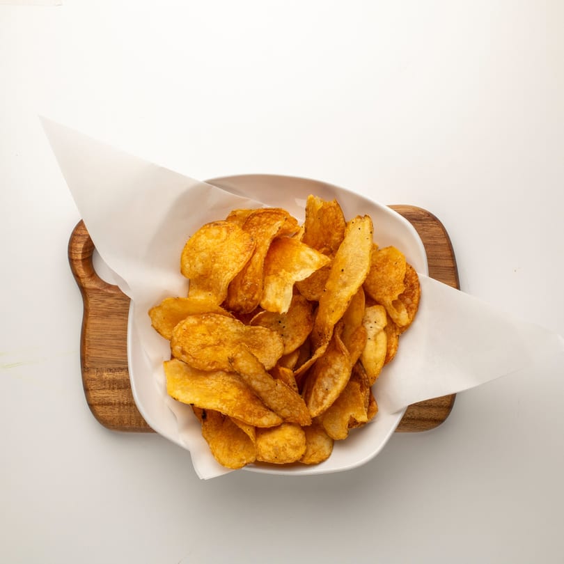 A white bowl filled with golden-brown, seasoned potato chips, lined with white parchment paper, on a wooden board.