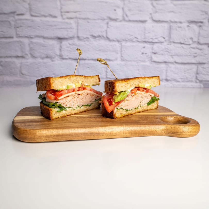 Two halves of a turkey sandwich with lettuce, tomato, avocado, and cheese on a wooden board.