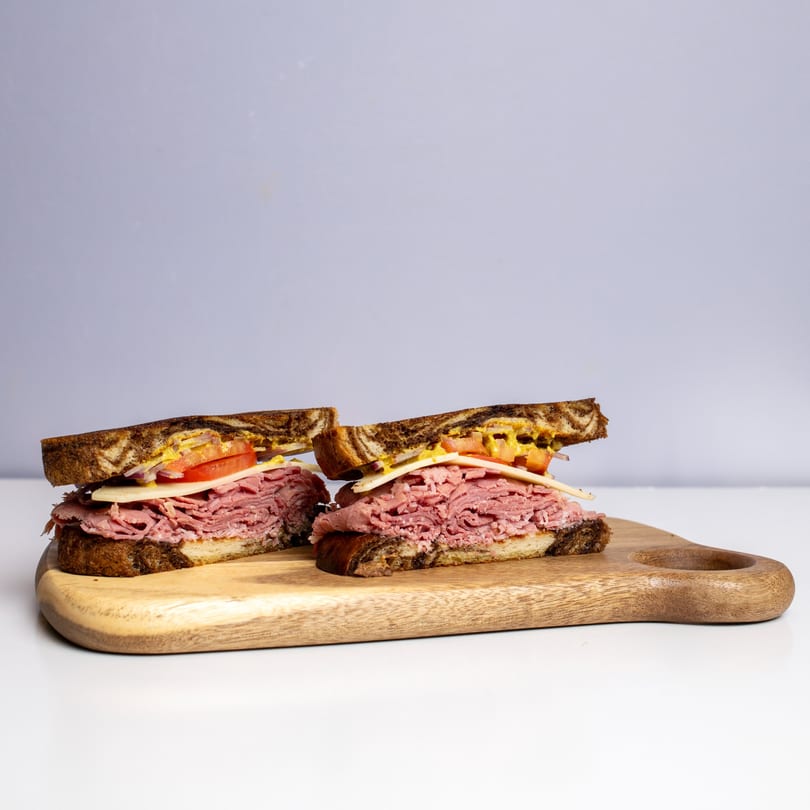 Two halves of a deli sandwich with pastrami, cheese, tomato, and mustard on rye bread.