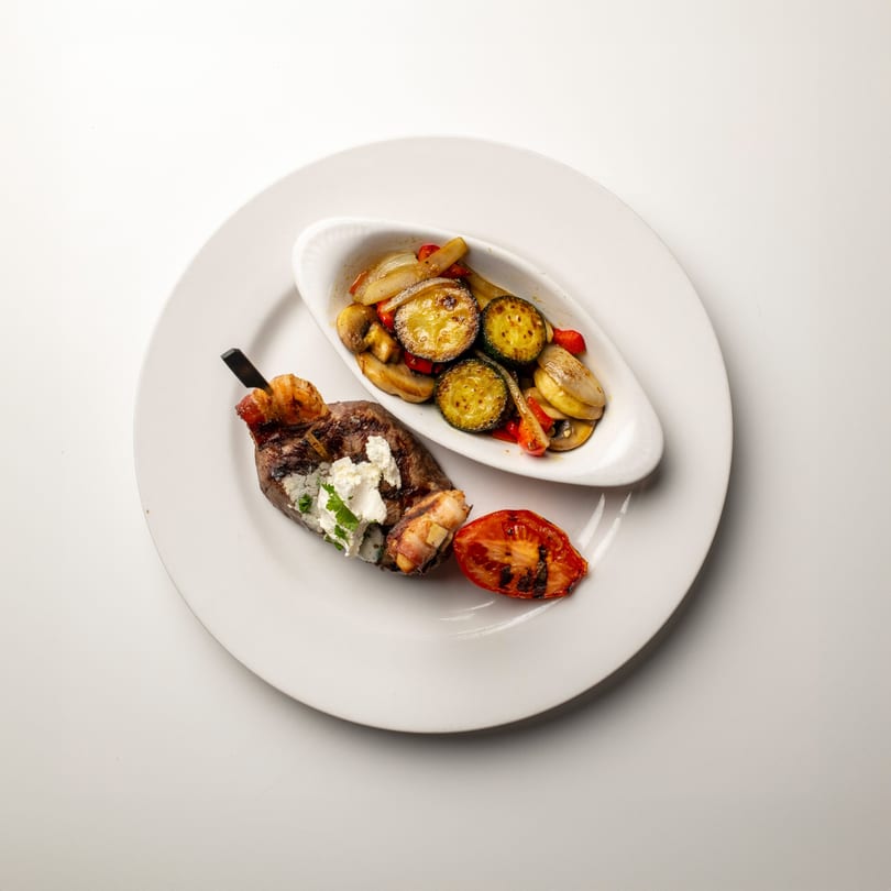 Bacon-wrapped steak with cream cheese, grilled tomato, and a side of roasted vegetables.