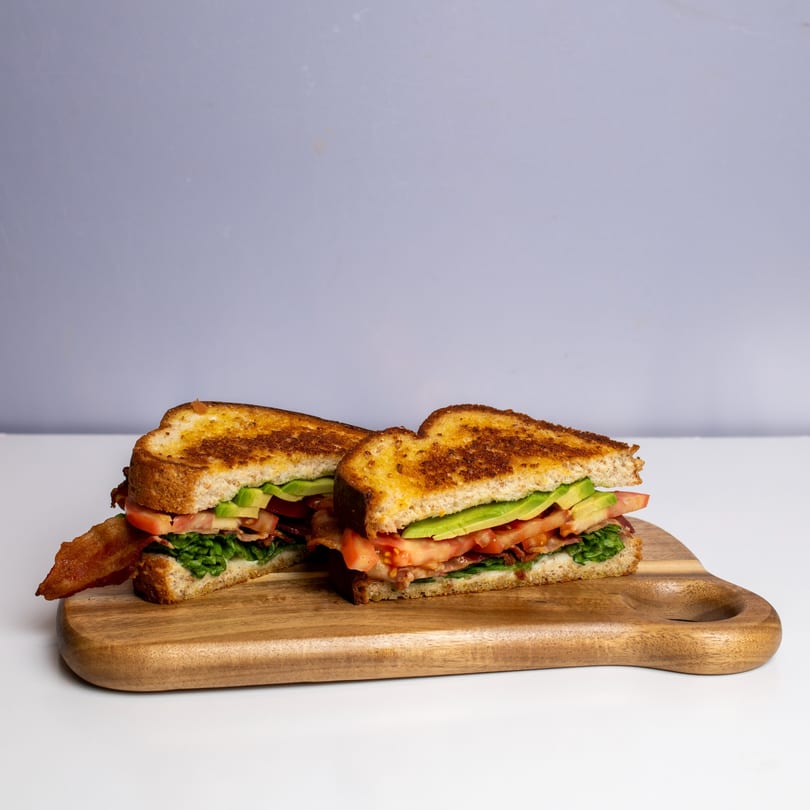 Two halves of a toasted BLT sandwich with avocado, bacon, lettuce, and tomato on a wooden board.