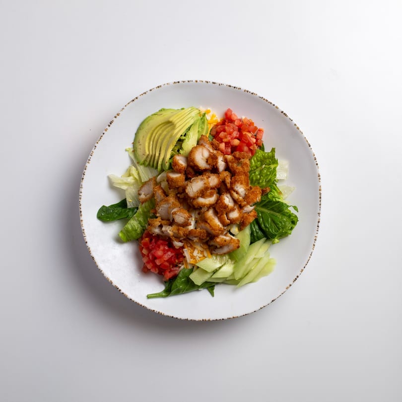 Crispy chicken salad with avocado, tomatoes, cucumbers, and mixed greens on a white plate.