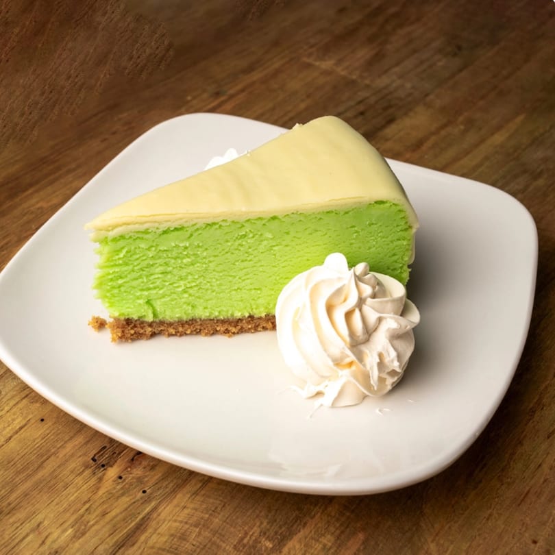 A slice of bright green cheesecake with a graham cracker crust and whipped cream on a white plate.