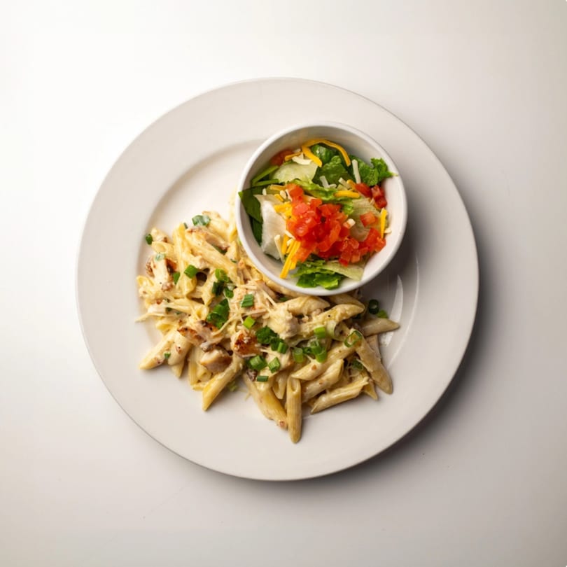 Creamy penne pasta with chicken and green onions, served with a side salad in a white bowl.