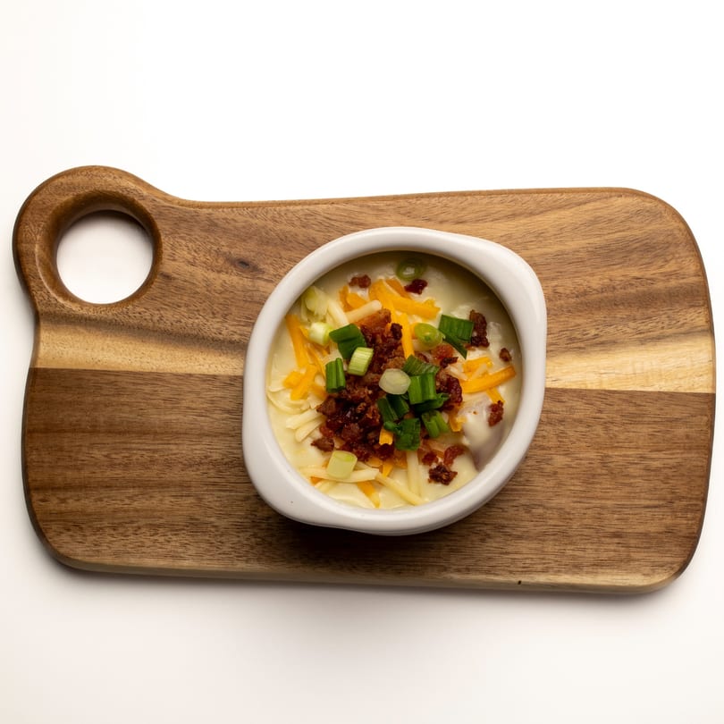 Creamy potato soup topped with shredded cheese, bacon bits, and green onions in a white bowl on a wooden board.