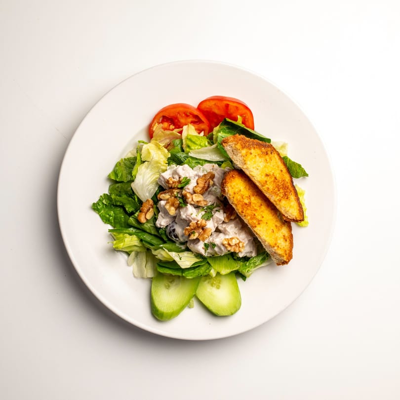 Chicken salad with walnuts, lettuce, tomatoes, cucumbers, and toasted bread on a white plate.