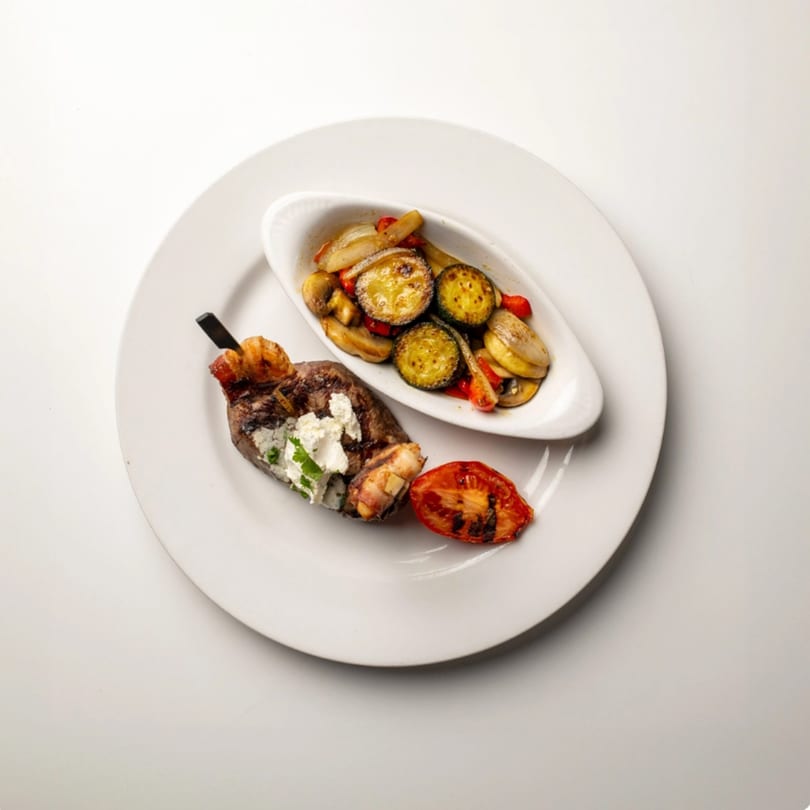 Bacon-wrapped steak with goat cheese, grilled tomato, and a side of roasted vegetables.