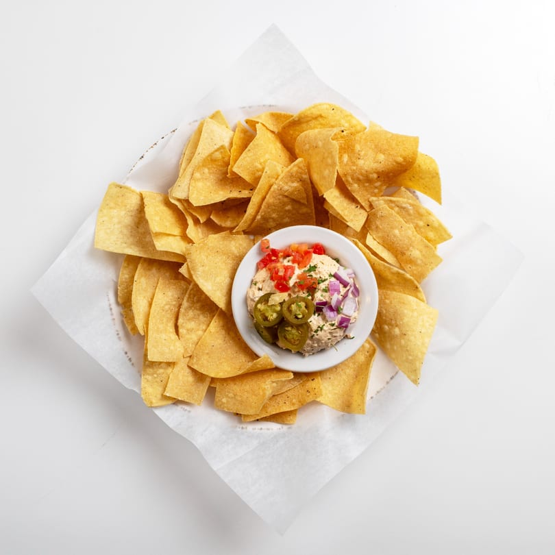 A basket of tortilla chips with a small bowl of dip topped with tomatoes, onions, and jalapeños.